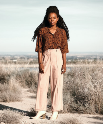 Modelo feminina vestindo blusa marrom, calça bege, par de sapatos altos na cor branca, cordão e pulseira, em um ambiente aberto com chão de terra e matos secos.