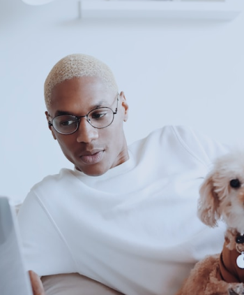 Modelo masculino com blusa branca e um par de óculos de lentes transparentes arredondadas, utilizando um notebook, com um cachorro poodle branco no seu colo, e em um fundo branco.